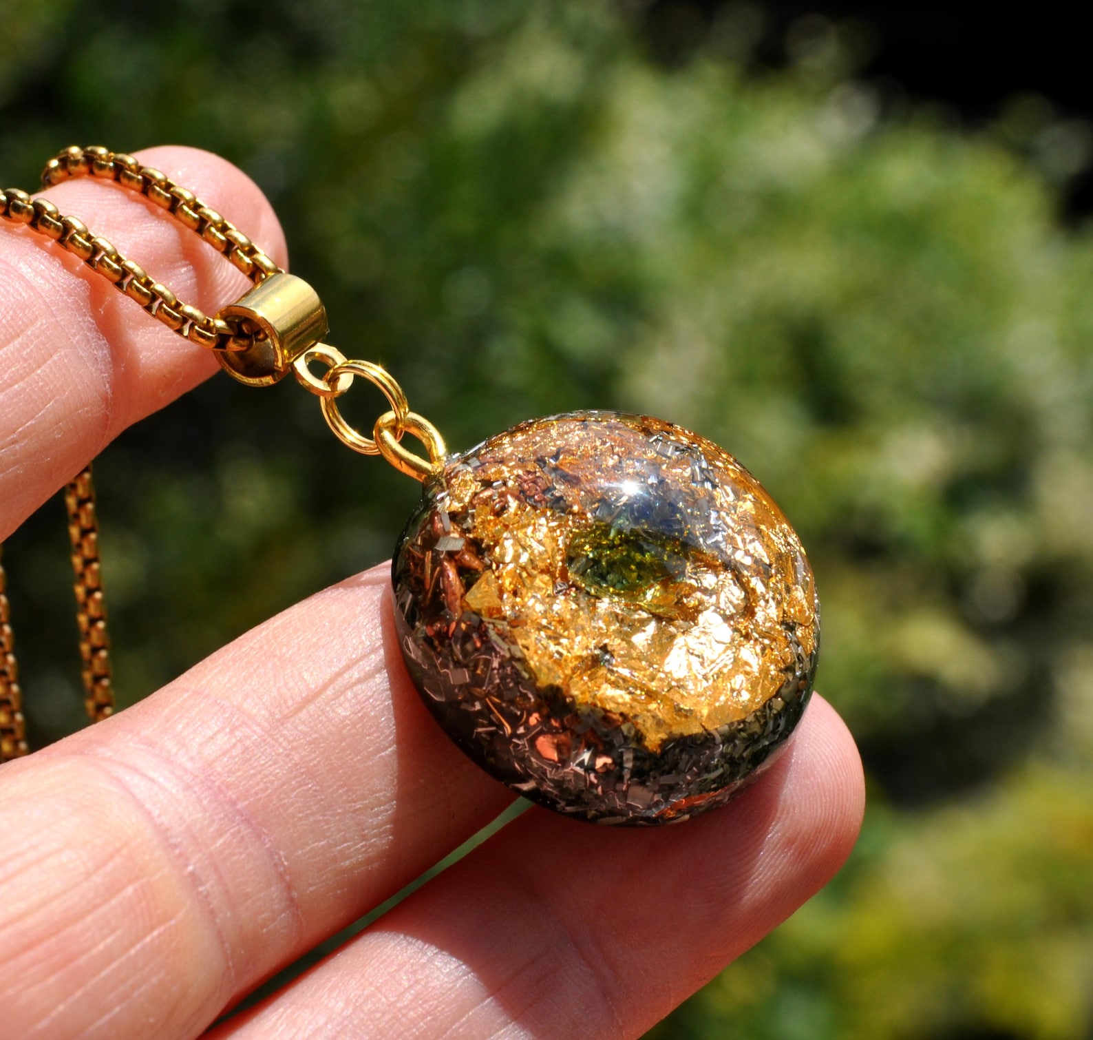 Small Moldavite Orgonite pendant with 24k gold flakes, held between fingers in natural sunlight.