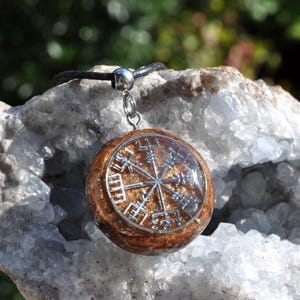 Op de afbeelding: Een ronde hanger met een zilverkleurig Vegvisir-symbool ingebed in een glinsterende, amberkleurige harsbasis. De hanger is met een zilverkleurig metalen oogje bevestigd aan een zwart koord, wat een contrast vormt tussen het metalen symbool en de glinsterende harsachtergrond.