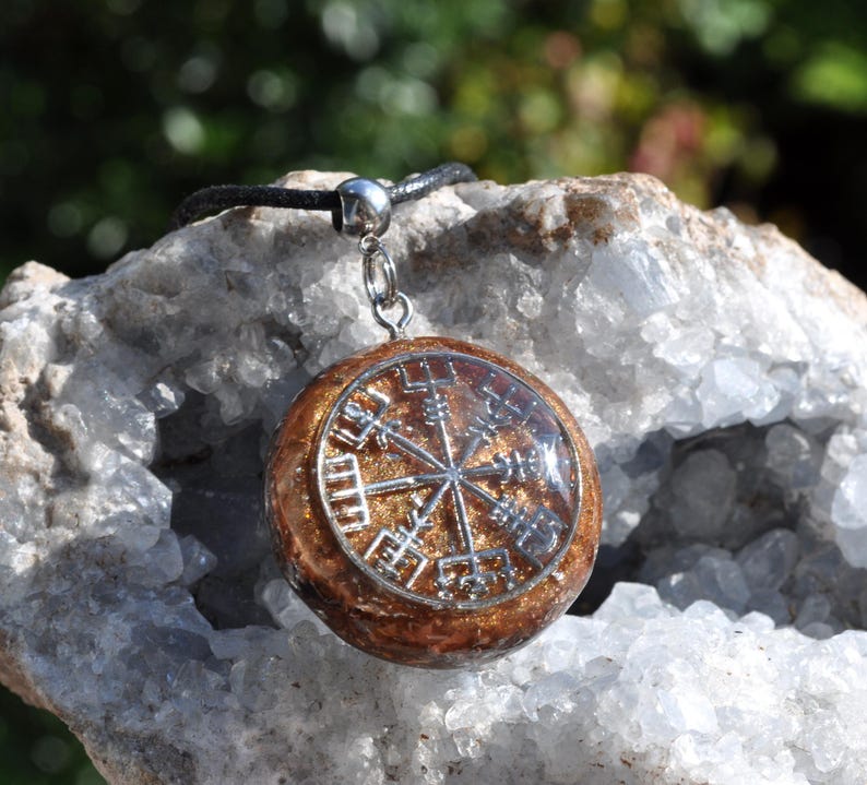 Op de afbeelding: Een ronde hanger met een zilverkleurig Vegvisir-symbool ingebed in een glinsterende, amberkleurige harsbasis. De hanger is met een zilverkleurig metalen oogje bevestigd aan een zwart koord, wat een contrast vormt tussen het metalen symbool en de glinsterende harsachtergrond.