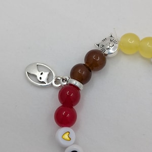 May include: A beaded bracelet with a cat charm and the letters "CA" spelled out. The bracelet features red, brown, and yellow beads, with a silver cat charm and a white bead with a yellow heart.
