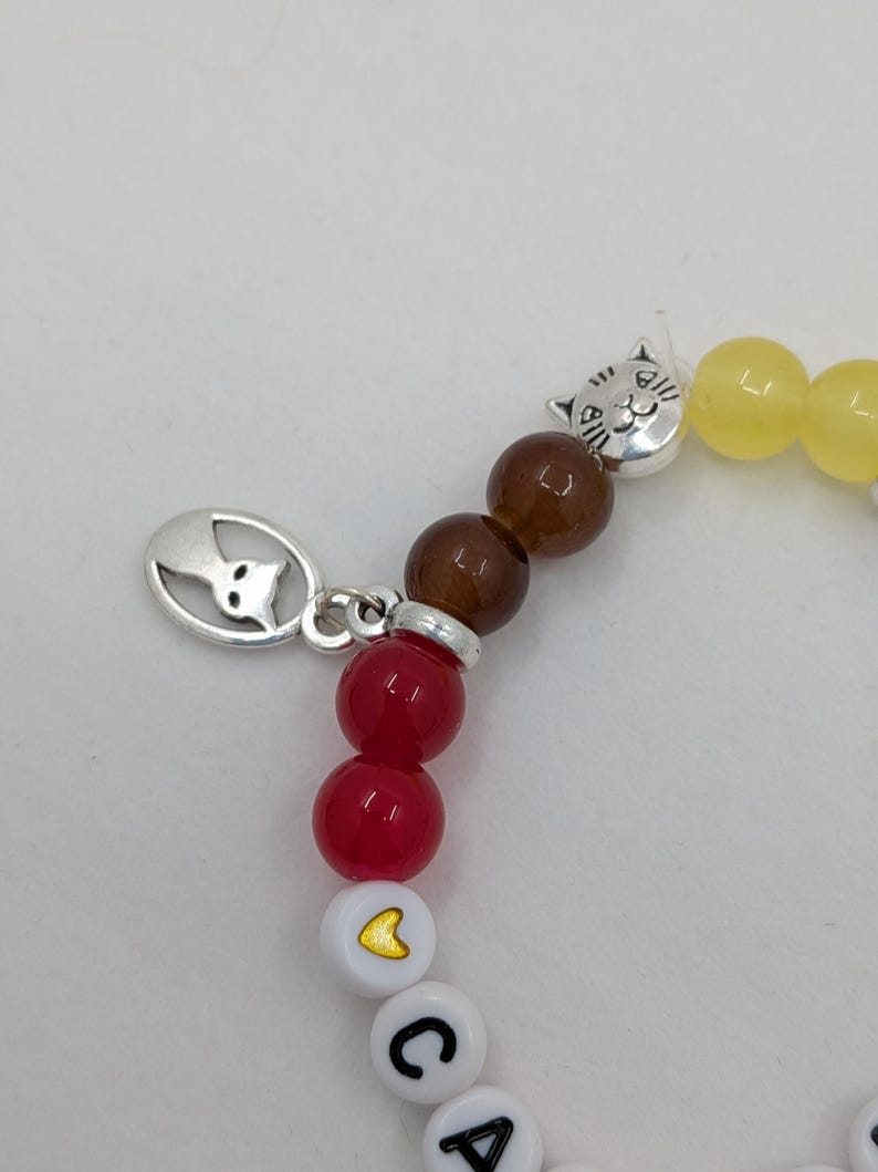 May include: A beaded bracelet with a cat charm and the letters "CA" spelled out. The bracelet features red, brown, and yellow beads, with a silver cat charm and a white bead with a yellow heart.