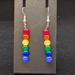 May include: A pair of earrings with a rainbow design. Each earring has seven round beads in red, orange, yellow, green, blue, and indigo. The beads are strung together with silver-coloured hooks.