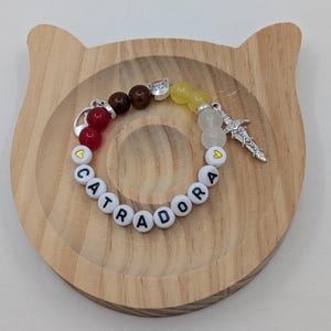 May include: A beaded bracelet with the word "CATRADORA" spelled out in white letter beads. The bracelet features red, brown, yellow, and clear beads, along with silver charms including a sword and a heart. The bracelet rests on a wooden cat-shaped tray.
