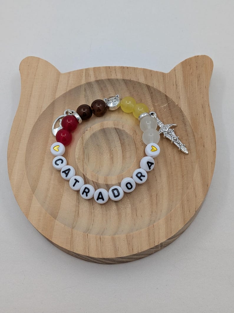 May include: A beaded bracelet with the word "CATRADORA" spelled out in white letter beads. The bracelet features red, brown, yellow, and clear beads, along with silver charms including a sword and a heart. The bracelet rests on a wooden cat-shaped tray.