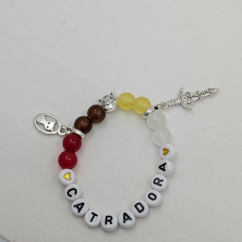 May include: A beaded bracelet with the word "CATRADORA" spelled out in white letter beads. The bracelet features red, brown, yellow, and white beads, along with silver charms of a cat and a sword.