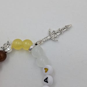 May include: A beaded bracelet featuring a silver sword charm and a cat bead. The bracelet includes brown, yellow, and white beads, with white letter beads spelling out a name. The bracelet is on a white background.