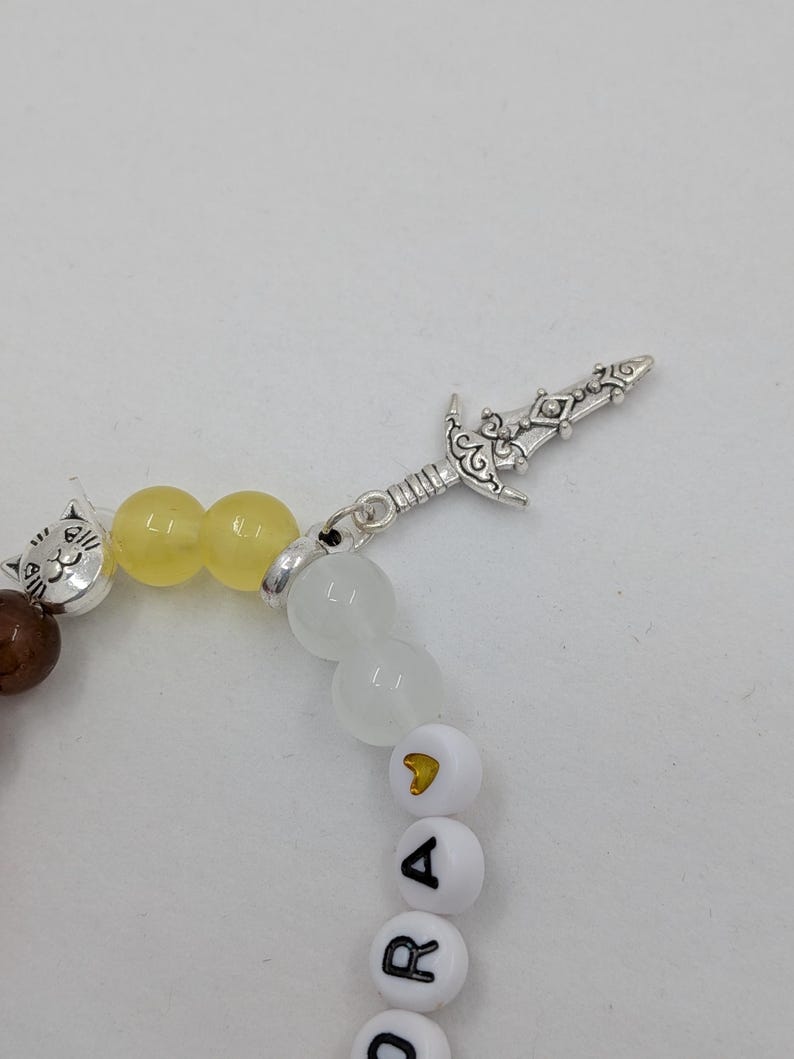 May include: A beaded bracelet featuring a silver sword charm and a cat bead. The bracelet includes brown, yellow, and white beads, with white letter beads spelling out a name. The bracelet is on a white background.
