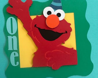 Puede incluir: Personaje rojo de Elmo con un gorro de fiesta a rayas azules y blancas, saludando con una gran sonrisa. El personaje está en un marco verde con un fondo amarillo. La palabra "one" está escrita en azul en el lado izquierdo de la imagen.