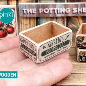 May include: A miniature wooden potting shed box with the text "MARTIN'S POTTING SHED" and "GARDEN TOOLS". The box is rectangular with a light brown interior and a white exterior with green text. The box is held in a hand.
