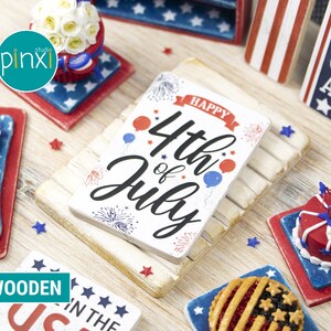 May include: A miniature wooden sign with the text "Happy 4th of July" in red, white, and blue. The sign is decorated with fireworks, stars, and balloons. The sign is on a white wooden surface with other miniature patriotic decorations.