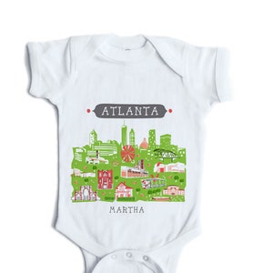 May include: White baby onesie with a green and red illustration of the Atlanta skyline. The text "ATLANTA" is in gray above the skyline and the text "MARTHA" is below the skyline.