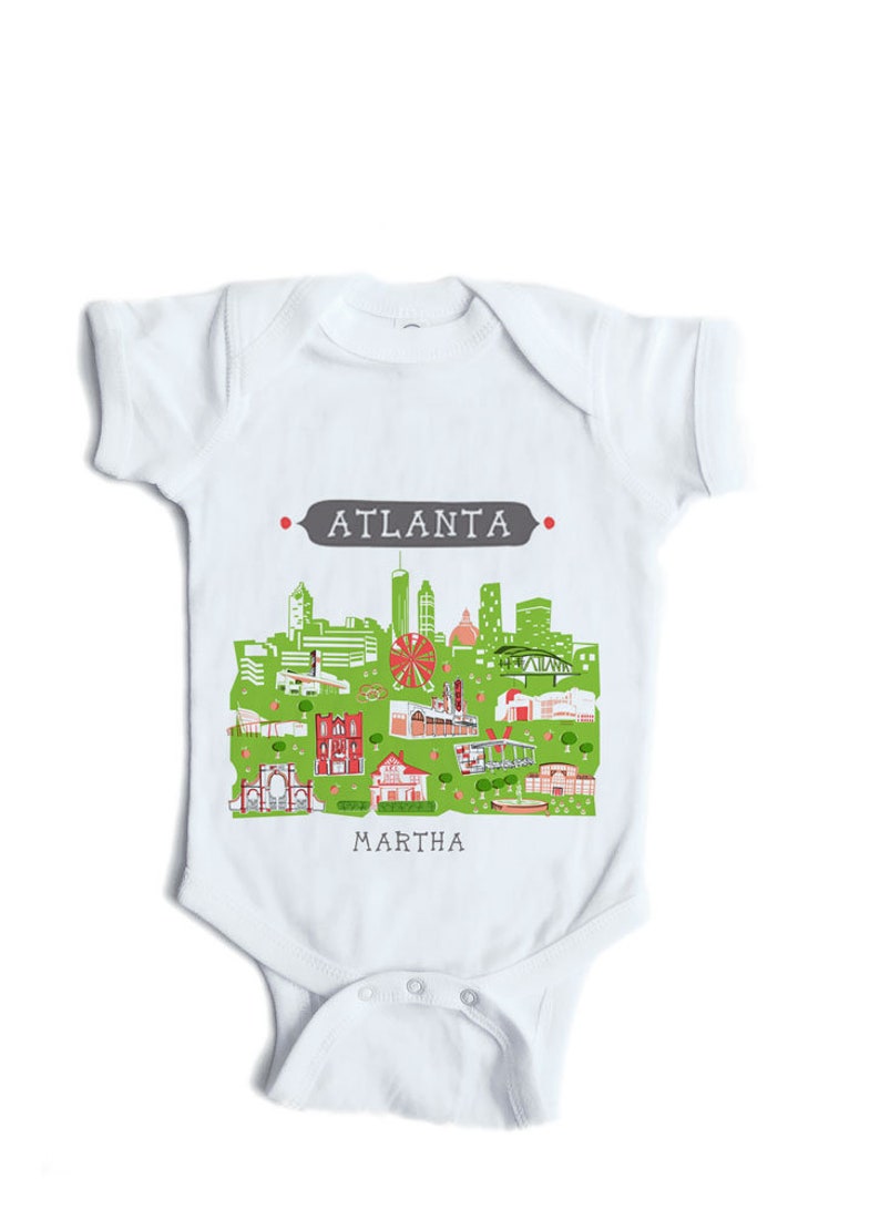 May include: White baby onesie with a green and red illustration of the Atlanta skyline. The text "ATLANTA" is in gray above the skyline and the text "MARTHA" is below the skyline.