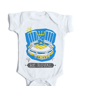 May include: White baby onesie with a blue and yellow illustration of a baseball stadium with the text "BE ROYAL" and a crown with the letters "KC" inside.