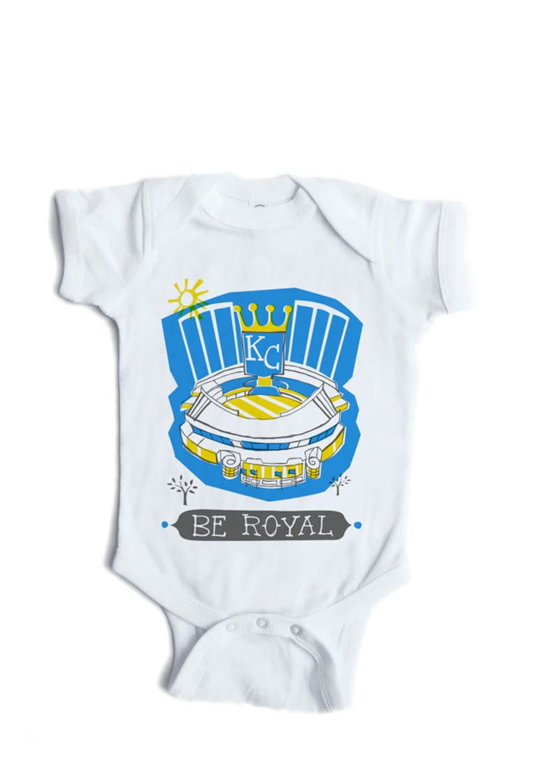May include: White baby onesie with a blue and yellow illustration of a baseball stadium with the text "BE ROYAL" and a crown with the letters "KC" inside.