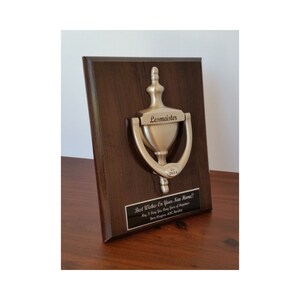 May include: A silver door knocker mounted on a dark wood plaque. The knocker is engraved with the name "Lesmeister" and the year "2014". A black plaque below the knocker reads "Best Wishes On Your New Home!! May It Bring You Many Years of Happiness. Ben Hagen, ABC Realty."