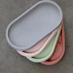 May include: A stack of four oval-shaped concrete trays in shades of gray, pink, green, and coral. The trays have a smooth surface and a raised edge. The trays are stacked on top of each other, with the gray tray on top.