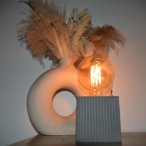 May include: A decorative arrangement featuring a cream-colored vase with a circular opening, dried pampas grass, and a vintage-style light bulb. The light bulb is set on a gray, corrugated metal base, creating a warm, inviting ambiance. The overall theme is modern home decor.