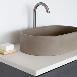 May include: A gray concrete oval vessel sink with a brushed nickel faucet mounted on a white countertop. A small wooden soap dish with a white bar of soap is on the countertop.