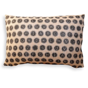 May include: Rectangular cushion with a beige burlap texture. The cushion features a pattern of black typewriter keys, each displaying a letter of the alphabet. A neutral home decor item.