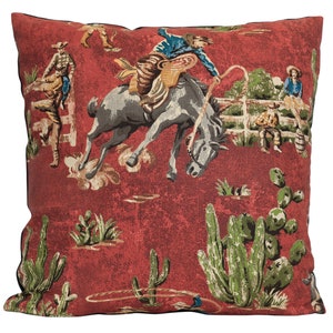 Puede incluir: Cojín rojo con un diseño vintage de vaquero y caballo. El cojín presenta un vaquero montando un caballo que se encabrita, cactus y otras imágenes del oeste.