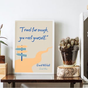 May include: A beige and orange print with a blue signpost and the quote "Travel far enough, you meet yourself." by David Mitchell. The print is framed in a white frame and is sitting on a wooden table.
