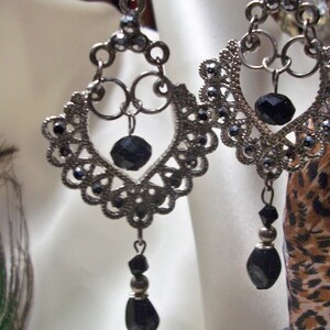 May include: A pair of silver dangle earrings with black beads. The earrings feature a delicate, intricate design with a heart shape at the center.