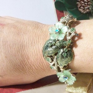 Pode incluir: Um bracelete vintage verde e dourado com um design floral. O bracelete apresenta um grupo de flores verdes com strass no centro. O bracelete &eacute; feito de metal e tem um visual delicado e feminino.