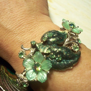 Pode incluir: Um bracelete de prata com strass verdes e transparentes e um design floral. O bracelete est&aacute; no pulso de uma pessoa.