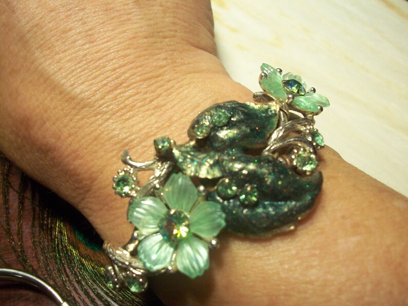 Pode incluir: Um bracelete de prata com strass verdes e transparentes e um design floral. O bracelete est&aacute; no pulso de uma pessoa.