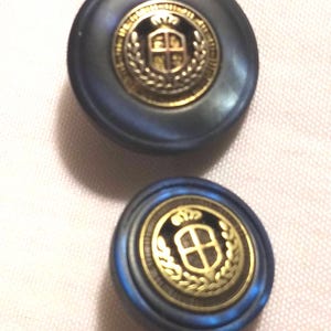 May include: Two blue and gold buttons with a crest design. The crest features a shield with a cross and a crown above it. The shield is surrounded by a wreath of leaves.