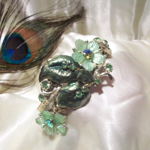 Pode incluir: Um broche floral prateado e verde com uma pena de pav&atilde;o no fundo. O broche apresenta um grupo de flores verdes com pedras de imita&ccedil;&atilde;o iridescentes no centro.