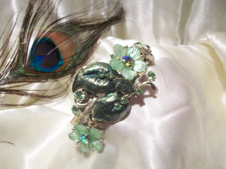 Pode incluir: Um broche floral prateado e verde com uma pena de pav&atilde;o no fundo. O broche apresenta um grupo de flores verdes com pedras de imita&ccedil;&atilde;o iridescentes no centro.