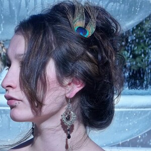 May include: A woman with dark brown hair styled in an updo with a peacock feather hair clip. She is wearing a silver dangle earring with black beads.