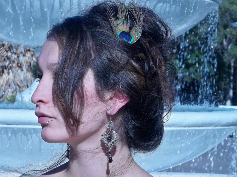 May include: A woman with dark brown hair styled in an updo with a peacock feather hair clip. She is wearing a silver dangle earring with black beads.
