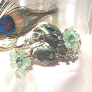 Pode incluir: Um bracelete de tom prateado com um design floral verde e prateado. O bracelete apresenta duas grandes flores verdes com pedras de imita&ccedil;&atilde;o iridiscentes no centro. As flores s&atilde;o rodeadas por flores verdes menores e pedras de imita&ccedil;&atilde;o.
