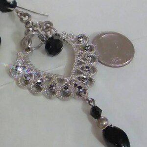 May include: Silver dangle earrings with black beads and a silver charm. The charm is shaped like a heart and has a filigree design. The earrings are accented with small, clear rhinestones.
