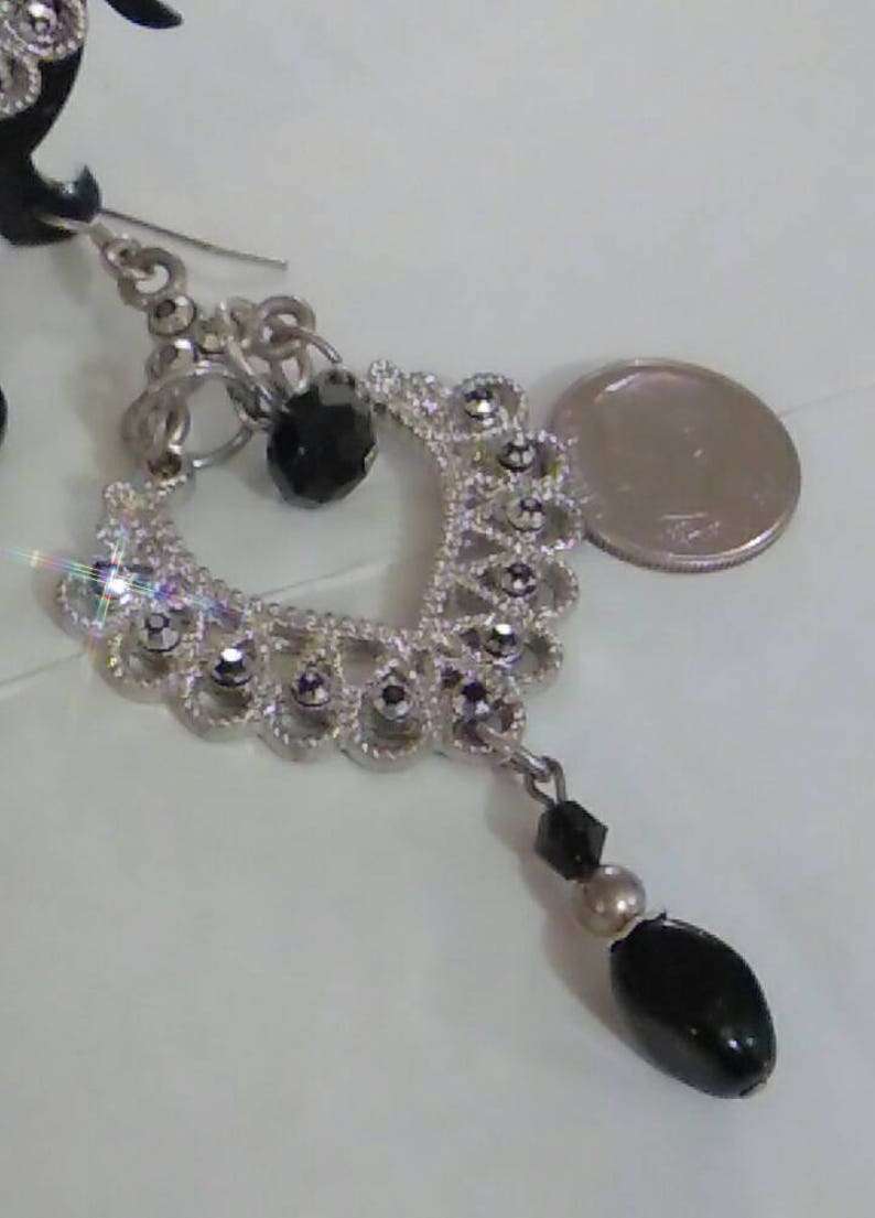May include: Silver dangle earrings with black beads and a silver charm. The charm is shaped like a heart and has a filigree design. The earrings are accented with small, clear rhinestones.