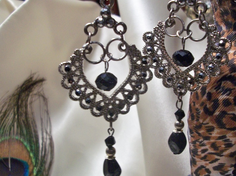 May include: A pair of silver dangle earrings with black beads. The earrings feature a delicate, intricate design with a heart shape at the center.