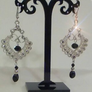 May include: A pair of silver dangle earrings with black beads. The earrings feature a delicate, intricate design with a heart shape at the center.