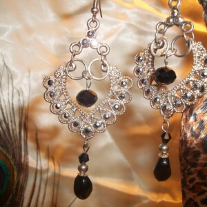 May include: Silver dangle earrings with black crystal accents. The earrings feature a delicate, intricate design with multiple layers of silver.