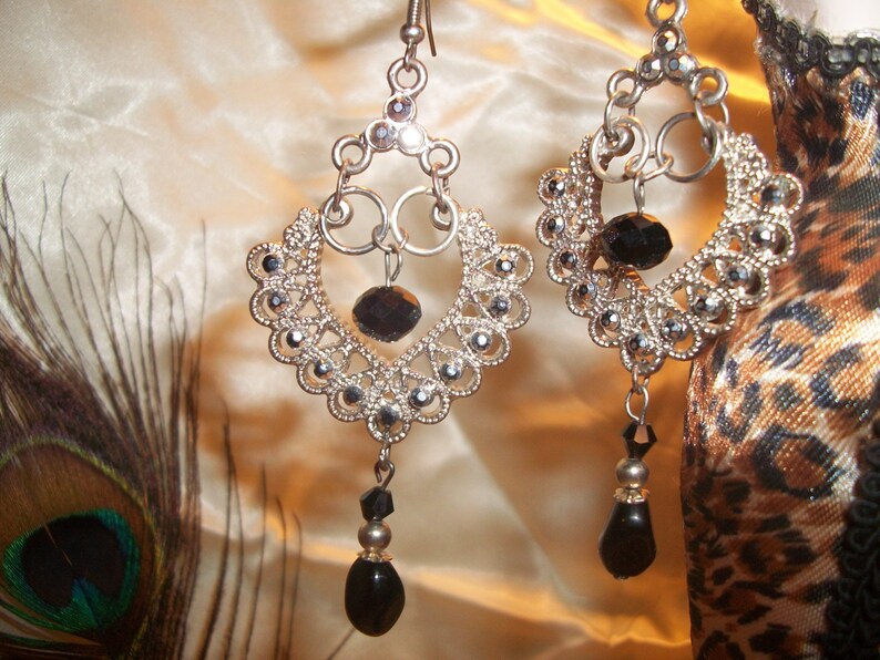 May include: Silver dangle earrings with black crystal accents. The earrings feature a delicate, intricate design with multiple layers of silver.