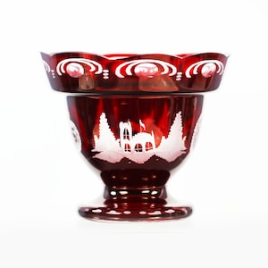 May include: A red glass bowl with a white etched design of a house and trees. The bowl has a scalloped rim and a footed base.