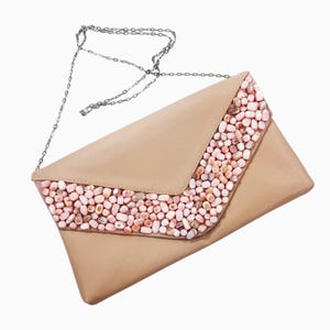 May include: A beige envelope-style clutch bag featuring a V-shaped flap adorned with numerous small, polished pink and white stone beads. The bag includes a thin, silver-toned metal chain strap attached to the top corners for carrying.