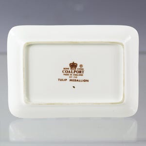 Puede incluir: Un plato rectangular de porcelana de color hueso con esquinas redondeadas. El plato presenta el texto "BONE CHINA COALPORT MADE IN ENGLAND EST 1750 TULIP MEDALLION" en letras marrones y un logotipo de corona.