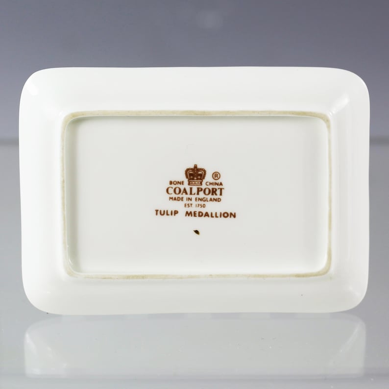 Puede incluir: Un plato rectangular de porcelana de color hueso con esquinas redondeadas. El plato presenta el texto "BONE CHINA COALPORT MADE IN ENGLAND EST 1750 TULIP MEDALLION" en letras marrones y un logotipo de corona.