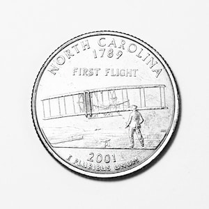 May include: A silver US quarter coin featuring the words "North Carolina 1789 First Flight" and the year "2001" along with the motto "E Pluribus Unum". The coin depicts a black and white image of the Wright brothers' first aeroplane.