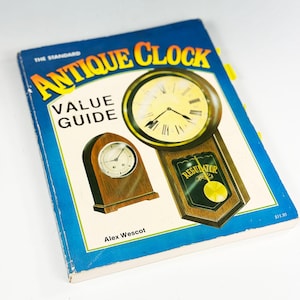 Può includere: Un libro vintage intitolato "The Standard Antique Clock Value Guide" con copertina blu e testo giallo. La copertina presenta illustrazioni di orologi antichi. Il nome dell'autore, Alex Wescot, è stampato in basso.