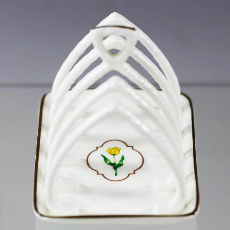 Puede incluir: Tostador de cer&aacute;mica blanca con asa y base rectangular. El soporte tiene separadores verticales y un emblema floral con un tulip&aacute;n amarillo. Los bordes est&aacute;n adornados con un borde marr&oacute;n.