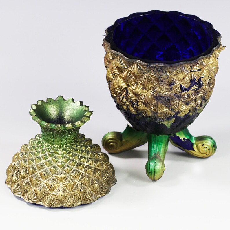 May include: A gold and green pineapple-shaped trinket box with a blue glass lid. The lid has a decorative pattern and the base has a textured surface.