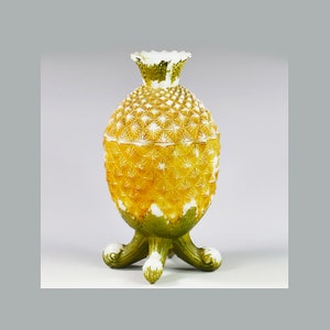Puede incluir: Un jarrón de vidrio en forma de piña amarillo y verde con una base blanca y tres patas verdes.